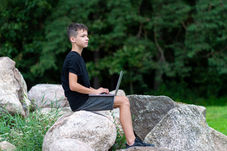 Boy sitting on a stone with laptop in garden. Kid have online web lesson or class on computer at nature, green class. Back to school. Outdoor learning conceptの写真素材