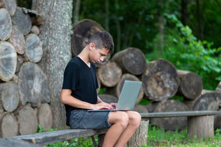 Boy sitting on wooden seat with laptop in forest. Kid have online web lesson or class on computer at nature, green class. Back to school. Outdoor learning conceptの写真素材