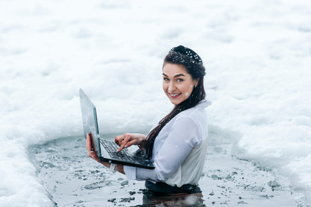 Girl with laptop in frozen lake ice hole. Woman hardening the body in cold water. Good immunity is protection against many diseases.の写真素材