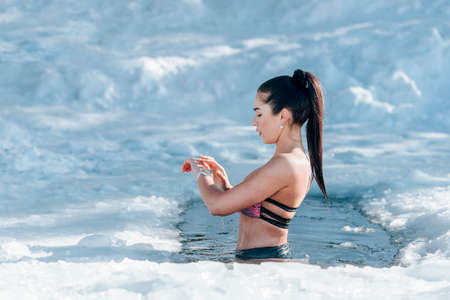 Girl with bikini and a watch in frozen lake ice hole. Woman hardening the body in cold water. Good immunity is protection against many diseases. Vintage color filterの写真素材