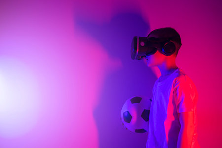 Young boy with soccer ball playing VR goggle on neon lighting. People, technology, sport and...