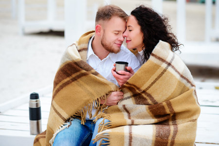 Young couple embracing and covered with a warm blanket sitting on autumn beach and drink coffeeの写真素材