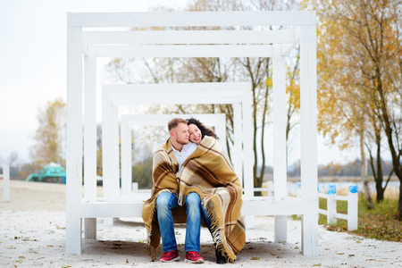 Young couple embracing and covered with a warm blanket sitting on autumn beachの写真素材