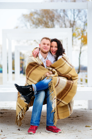 Young couple embracing and covered with a warm blanket sitting on autumn beachの写真素材