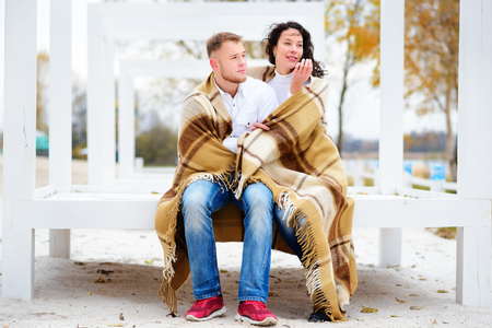 Young couple embracing and covered with a warm blanket sitting on autumn beachの写真素材