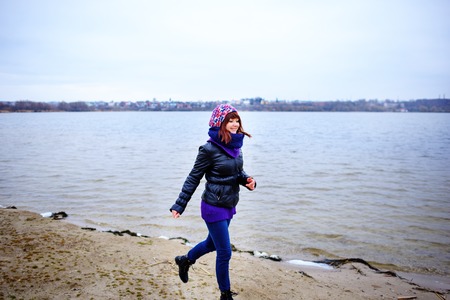 Lifestyle portrait of young Caucasian slim woman with messy long hair wearing black jeans and red shirt walking on windy day outdoor on beach shore at sunset, motion action blurの写真素材