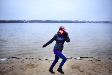 Lifestyle portrait of young Caucasian slim woman with messy long hair wearing black jeans and red shirt walking on windy day outdoor on beach shore at sunset, motion action blurの写真素材