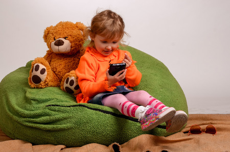 little girl sits on a high green pillow and plays with mobile phoneの写真素材