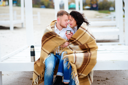 Young couple embracing and covered with a warm blanket sitting on autumn beach and drink coffee from a thermosの写真素材