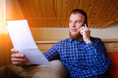 Serious mature man holding paper and talking on the mobile phone while sitting on the couch at homeの写真素材