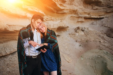 Beautiful woman and handsome man wrapped in a blanket. They are smiling and looking at the screen of a tablet on the background of a sand quarry.の写真素材