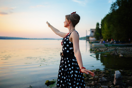 A beautiful young woman dressed standing by the lake, on the background of the sunset.の写真素材