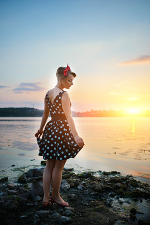 A beautiful young woman dressed standing by the lake, on the background of the sunset.の写真素材