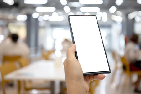 Smartphone with blank screen held in hand at food court, Restaurant in a shopping mall, Blurred background with people, Perfect for mobile app or banner website content placementの写真素材