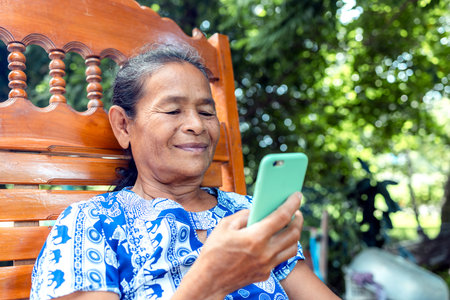 happy asian senior mature woman relaxing with video and face call, speaking on mobile phone with family, thai asia elderly grandmother enjoy smart outdoor lifestyle with wireless technology deviceの写真素材