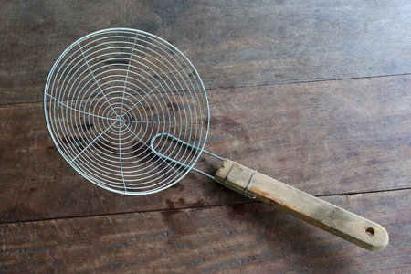 Metal wire skimmer with wooden handle on a rustic wooden table. Traditional kitchen utensil for...
