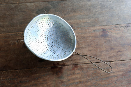 Traditional metal colander with perforated holes on a rustic wooden table. Kitchen utensil for draining, rinsing food, home cooking and culinary useの写真素材