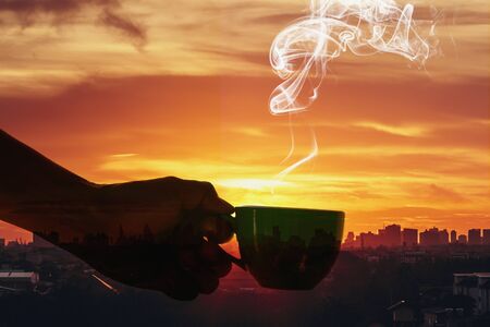 Man hand holding a cup of hot coffee aroma drifting far on the background silhouette of the city with sunrise,Concept  Double exposureの写真素材