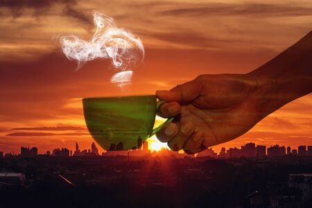 Man hand holding a cup of hot coffee aroma drifting far on the background silhouette of the city with sunrise,Concept  Double exposureの写真素材