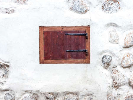 Closed window with shutters on the old style metal hinges. White uneven wall with fragments of stones visibleの写真素材