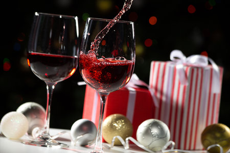 Beautiful splash of red wine in crystal glasses on the background of Christmas lights and Christmas gifts and ballsの写真素材