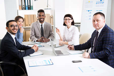 Multy-ethnic group of business people working while sitting at the office desk and smiling at cameraの写真素材