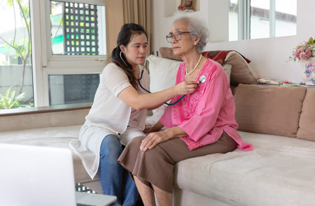 Asian female nurse checking body by stethoscope,senior woman sitting on sofa at homeの写真素材