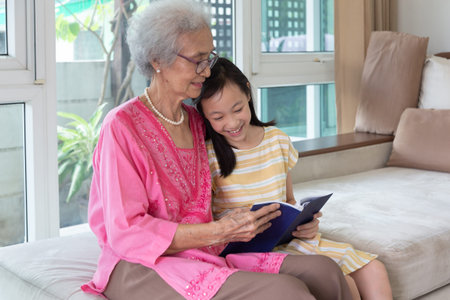 Grandmother and granddaughter sitting on sofa and reading book happy together at homeの写真素材