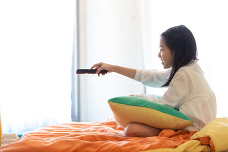 Asian girl watching TV lying on bed with remote control in handの写真素材