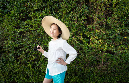 Outdoor portrait of a pretty asian girl in hat holding sunglasses,closeup of beautiful girl against greenery garden wallの写真素材