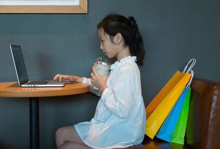 Asian girl drinking while keyboarding a new article using laptop computer sitting at a...