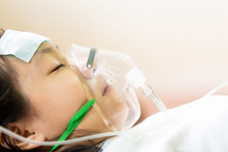 Asian little girl with oxygen mask and cold gel placed on her forehead to relief fever,cute child patient putting inhalation in hospital bed or home,intensive care,health care,support,help conceptの写真素材