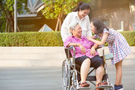 Asian senior woman having happiness and smiling with her daughter and granddaughter on wheelchair at outdoor park,elderly woman is happy with their family,child girl having fun talk,laughing together,happy family conceptの写真素材