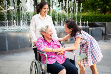 Asian senior woman having happiness and smiling with her daughter and granddaughter on wheelchair at outdoor park,elderly woman is happy with their family,child girl having fun talk,laughing together,happy family conceptの写真素材