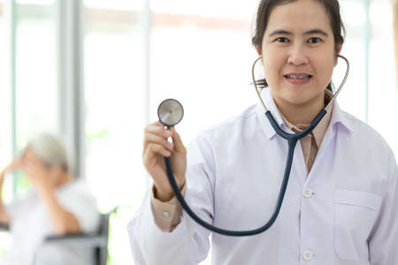 Asian female medical doctor holding stethoscope or young nurse standing,smiling and looking at camera,senior people sitting in wheelchair as a background,health care conceptの写真素材