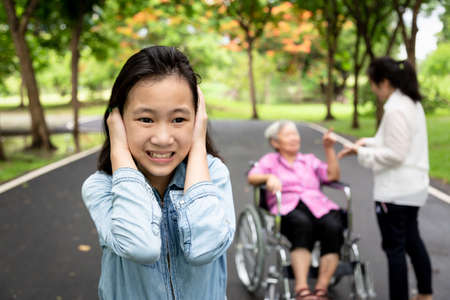 Asian daughter closed ears with hands, little child girl did not want to hear parents,grandmother in wheelchair and mother arguing,quarreling in outdoor park,problems in multi generation family,quarrel conceptの写真素材
