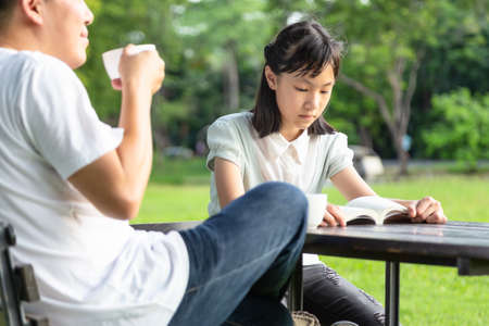Happy asian little child girl or daughter reading a book relax in outdoor park,enjoy,talk together,father holding cup hot drink coffee or tea in his hand in the morning in summer green nature,family,leisure,relationship conceptの写真素材