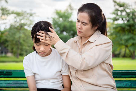 Asian beautiful girl has discomfort and headache,mother touching forehead of little cute child girl,checking the temperature,caring mom takes care of her sick daughter,high fever symptoms at outdoor park,medical and health care conceptの写真素材