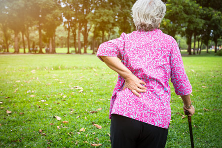 Asian senior woman hands touching back pain while walking in nature in summer,female patient having...