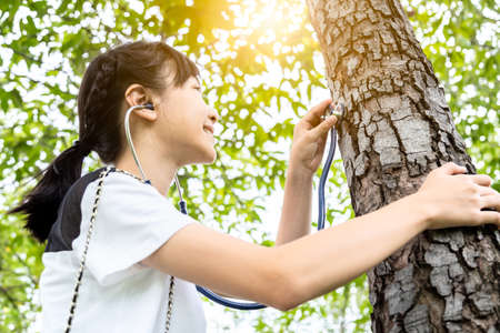 Asian child girl hand listening and care of a tree with stethoscope,dream job,female teenager want to be a doctor,checking healthy green trees in forest,love nature,protect environment,ecology conceptの写真素材