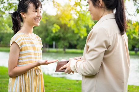 Asian woman hands open wallet,mother or guardian giving pocket money to daughter,smiling beautiful child girl demanding money, allowance,parent pulls out money from wallet to give her in outdoor parkの写真素材