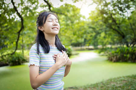 Happy smiling asian child girl standing in green nature,holding hands on heart,female teenage touching her chest,enjoy breathing fresh air with closed eyes,relaxing in park,healthy lifestyle conceptの写真素材