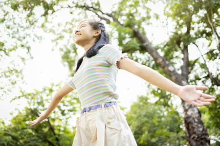 Happy smiling asian child girl standing with arms outstretched in green nature,female teenage enjoy breathing fresh air with raised arms up and closed eyes having fun,relax in park,healthy lifestyleの写真素材