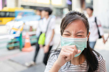 Asian child girl suffer from cough with face mask protection,sick female teen wearing medical mask because of air pollution in the city street,dust flies in the air,concept of pollution,dust allergiesの写真素材