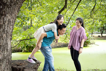 Happy asian family,father laugh giving piggyback ride to beautiful daughter having fun together, smiling mother enjoying active game with family, man carrying child girl on back, play in outdoor parkの写真素材