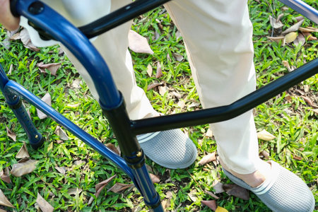Senior asian female patient trying to walk use walking aid during rehabilitation, elderly woman walking alone with walker for safety, practicing walk on the grass at park, disability concept,Closeupの写真素材