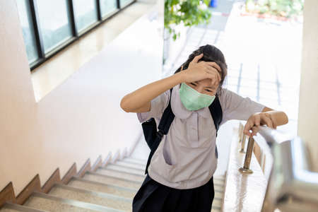 Tired asian child girl suffering from headache migraine,touches head or forehead with hand while walking up the stairs at school,teen student with a mask has flu,headache from stress,illness and feverの写真素材