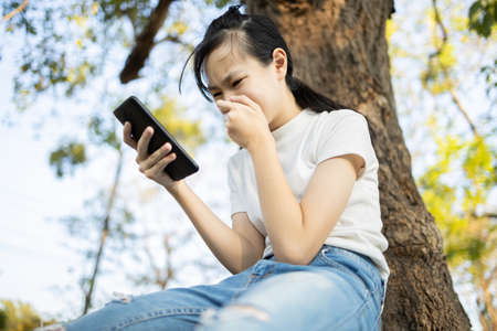Sad asian student victim of cyber bullying online sitting alone outdoor at school,teenage girl receiving text message at mobile phone, abuse cyberbullying being stalked,harassed,scared and desperate の写真素材