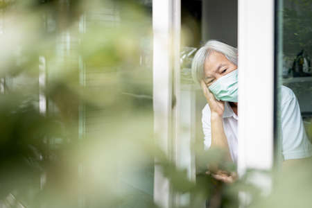Bored senior woman sit alone at home,elderly is looking outside through window,feeling bored,depressed and sad, unhappy old people after self-quarantine,stay home during Covid-19,Coronavirus pandemicの写真素材