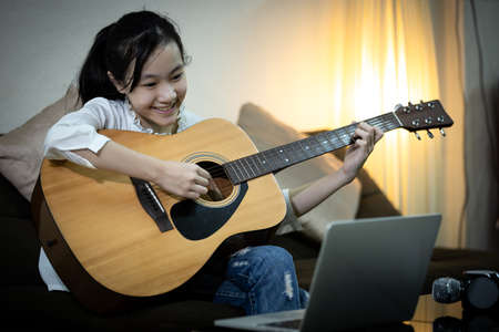 Happy smiling asian child girl is singing,playing the guitar to enjoy,fun,student is studying tutor with teacher,video conference calling on laptop computer,learn teach in online,distance teachingの写真素材